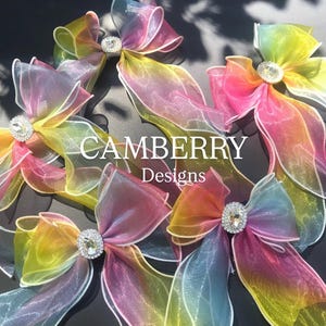 May include: Several colorful, sheer fabric bows with a gradient of colors, including pink, yellow, and blue. Each bow has a sparkling, round, rhinestone center. The text "CAMBERRY Designs" is visible.