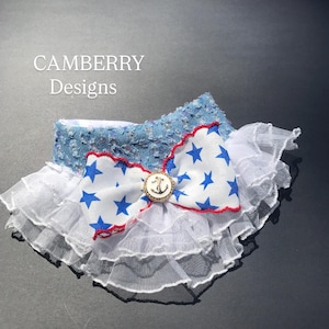 May include: A pet collar featuring a blue denim band with silver sequins, a white ruffled tulle trim, and a central white bow with blue stars, a red stitched edge, and a gold-toned circular anchor emblem. The text CAMBERRY Designs is visible.