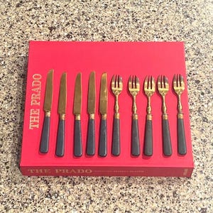 May include: A set of gold-toned knives and forks with dark gray handles arranged on a red box. The box has the text "THE PRADO" in gold lettering. The flatware is displayed on a speckled surface.