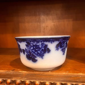 May include: A white and blue porcelain bowl with a floral design. The bowl features a rounded shape with a flared rim. The blue floral pattern is set against a white background. The bowl is sitting on a wooden surface.