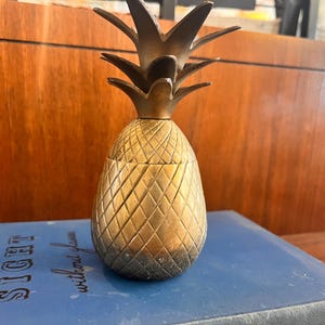 May include: A brass pineapple-shaped container with a textured surface and a removable top. The container sits on a blue book with the words "SIGHT without glasses" visible. The pineapple has a detailed, diamond-patterned design.
