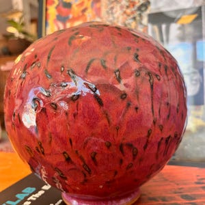 May include: A spherical ceramic vase with a glossy finish, featuring a vibrant red and magenta base color with dark, vertical drip-like markings. The surface has a textured, dimpled appearance, and the vase sits on a small, circular base.