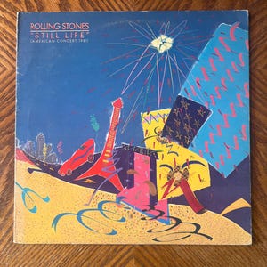 May include: A square vinyl record cover for The Rolling Stones Still Life American Concert 1981. The blue background features a colorful, abstract illustration with a red guitar, yellow shapes, and fireworks, with the title text in white.