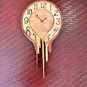 May include: A round wooden wall clock with a natural light wood finish and a live edge border. The clock face features black Arabic numerals from 1 to 12, with a melting or dripping design extending downward from the bottom of the circular frame.