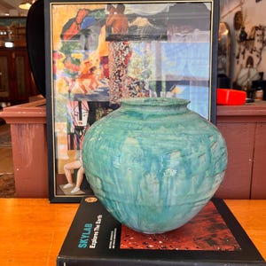 May include: A round, teal-colored ceramic vase with a glossy, marbled glaze and horizontal ridges, resting on a black book titled SKYLAB Explores The Earth, positioned in front of a framed collage of various artistic images and figures.