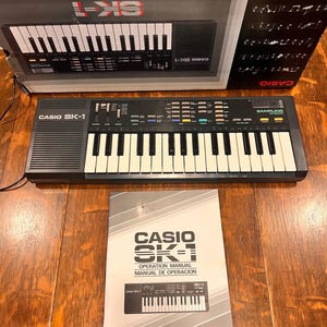 May include: A black Casio SK-1 keyboard with white keys and a control panel. The keyboard is displayed with its original box and an operation manual. The box and manual feature the Casio SK-1 logo and text.