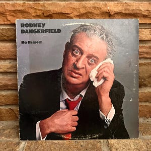 May include: Album cover for Rodney Dangerfield's "No Respect." The cover features a portrait of Dangerfield in a suit and tie, holding a handkerchief to his face. The album title and artist's name are printed at the top.