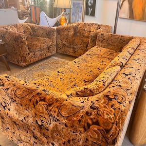 May include: A living room furniture set consisting of a long sofa and two matching armchairs. Each piece is upholstered in a plush, velvet-like fabric featuring a prominent, dark brown and golden-orange swirling paisley pattern throughout.