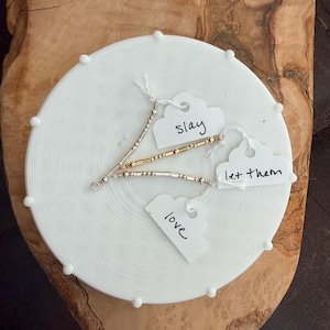 Puede incluir: Tres pulseras de cuentas con etiquetas que dicen "slay", "let them" y "love" se exhiben sobre una superficie circular blanca y texturizada. Las pulseras tienen cuentas plateadas y doradas. El fondo es una superficie de madera.