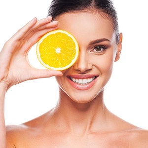 May include: A woman smiles while holding a slice of orange in front of her eye.