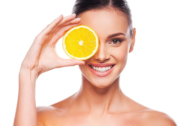 May include: A woman smiles while holding a slice of orange in front of her eye.
