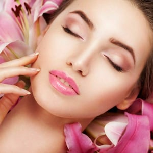 May include: A woman with closed eyes and pink lipstick lies on a bed of pink lily petals.