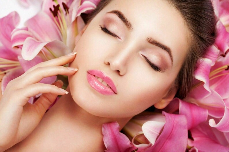 May include: A woman with closed eyes and pink lipstick lies on a bed of pink lily petals.