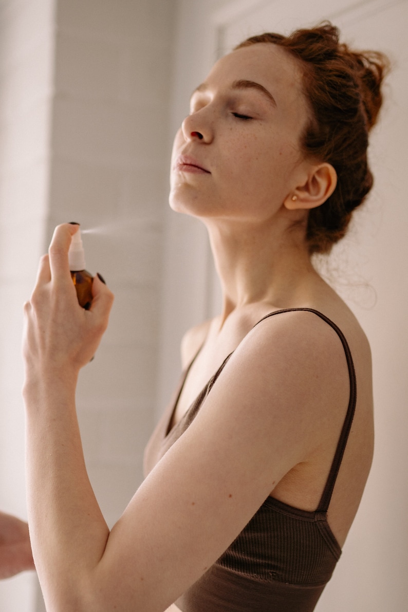 May include: A woman with red hair is spraying a clear bottle of facial mist on her face. She is wearing a brown tank top.