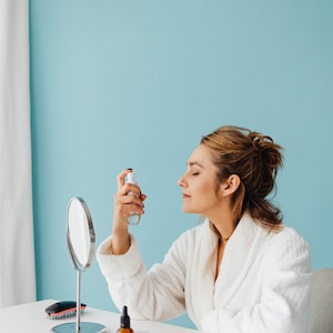 May include: A woman in a white bathrobe is spraying a facial mist in front of a mirror.  A small bottle of serum and a hairbrush are on the table in front of her.
