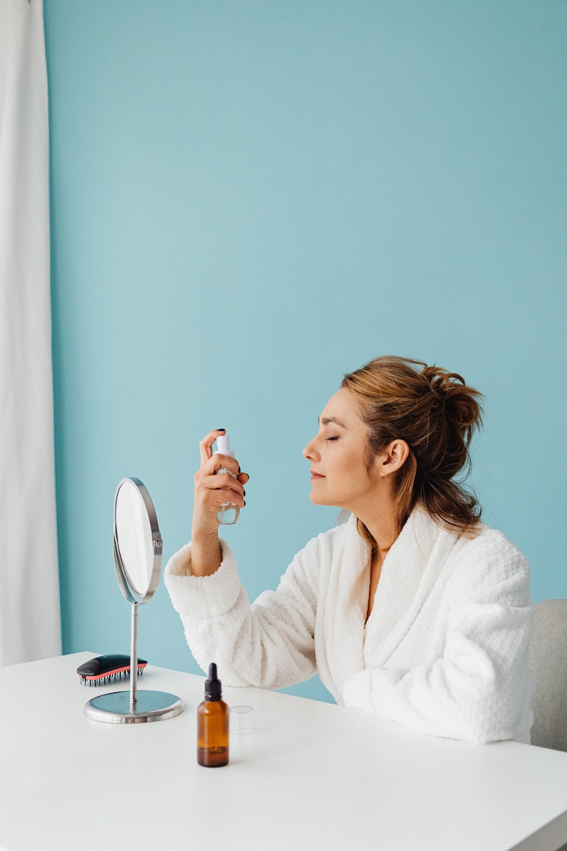 May include: A woman in a white bathrobe is spraying a facial mist in front of a mirror.  A small bottle of serum and a hairbrush are on the table in front of her.