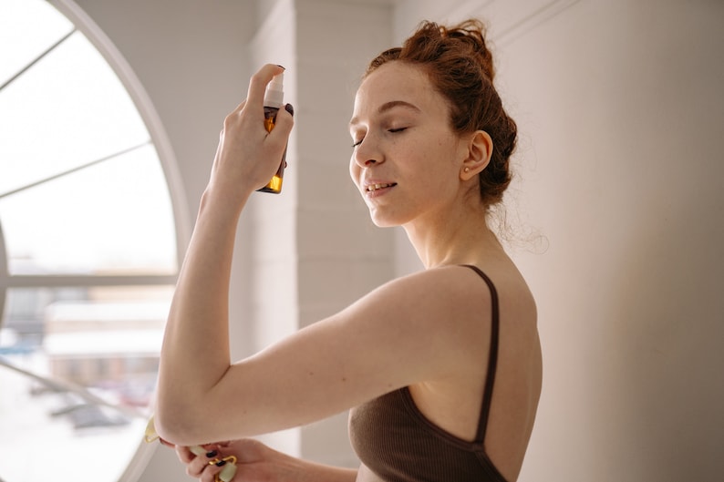 May include: A woman with red hair sprays a brown bottle of facial mist on her face. She is wearing a brown tank top.