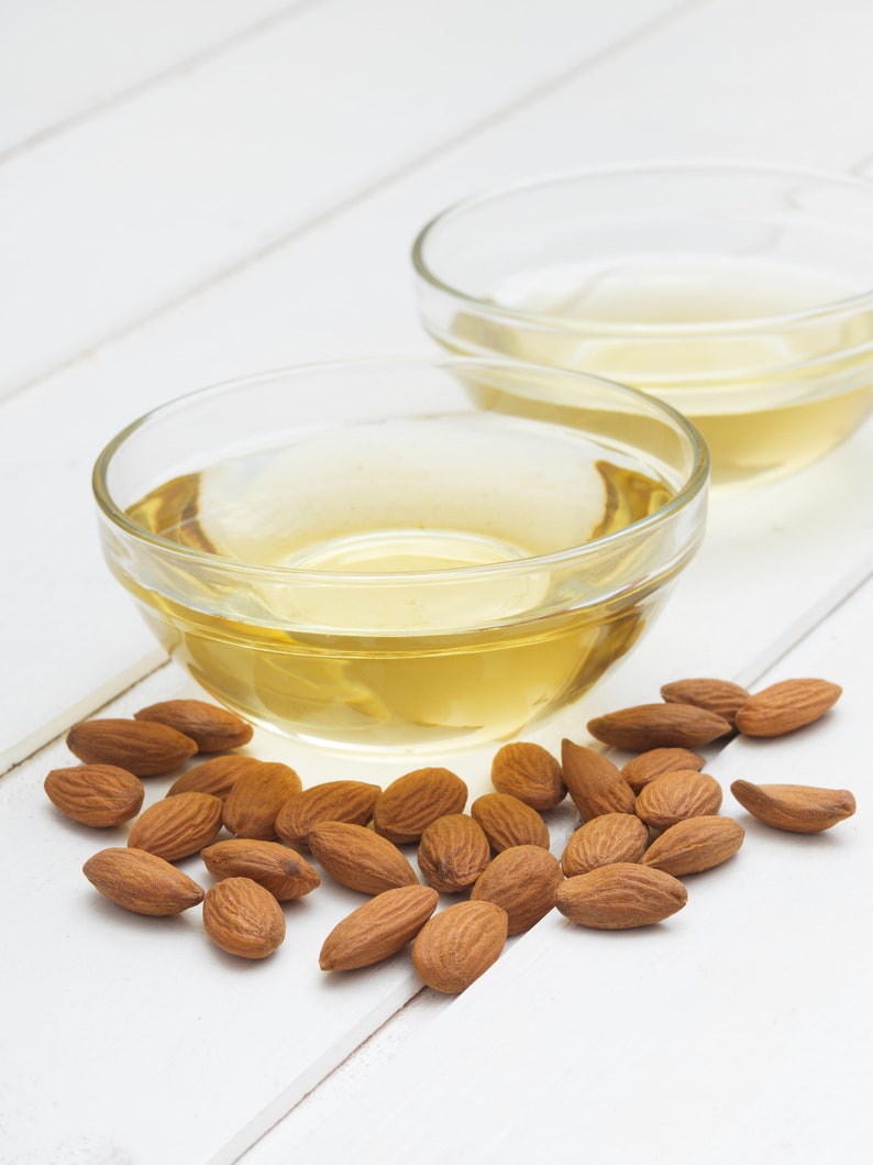 May include: Two clear glass bowls filled with light yellow almond oil, surrounded by a scattering of brown almonds on a white wooden surface.