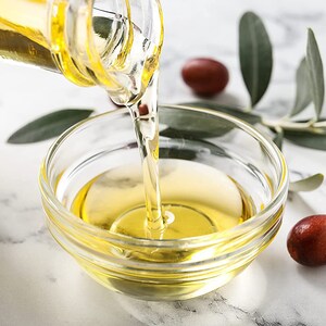 May include: A clear glass bottle pouring golden yellow oil into a glass bowl. The bowl is sitting on a white marble surface. There are red berries and green leaves in the background.