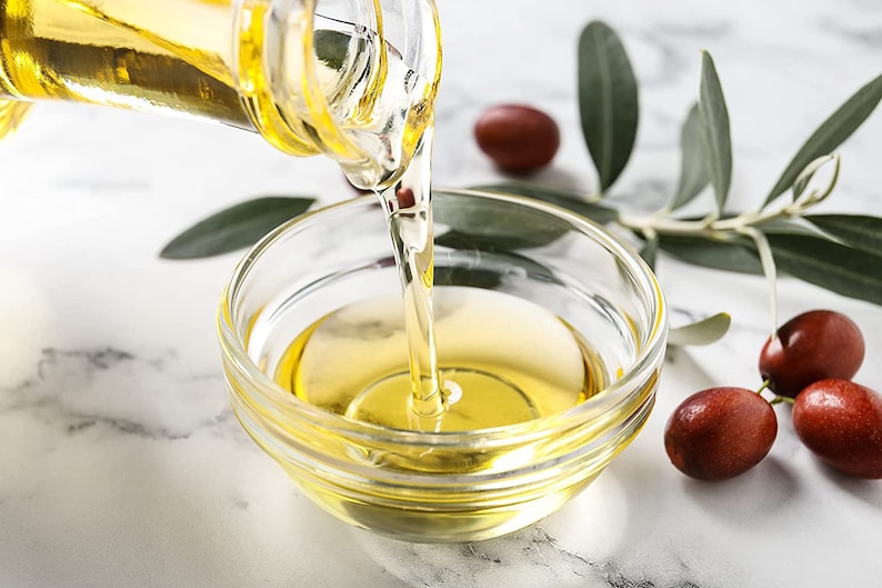 May include: A clear glass bottle pouring golden yellow oil into a glass bowl. The bowl is sitting on a white marble surface. There are red berries and green leaves in the background.