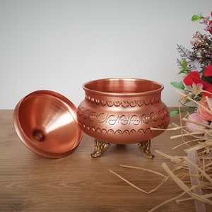 May include: A copper sugar bowl with a lid and decorative engravings. The bowl has four feet and is sitting on a wooden surface.