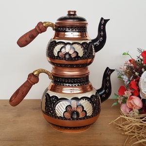 May include: A traditional Turkish double-spout copper teapot with black and white floral designs. The teapot has two wooden handles and a copper lid.
