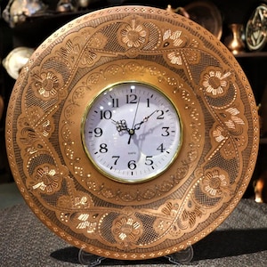May include: A round, copper-colored wall clock with an ornate floral and scroll design. The clock face is white with black numerals and hands. The clock is approximately 12 inches in diameter, showcasing a vintage aesthetic.
