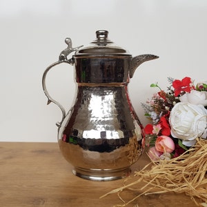 May include: A silver hammered metal pitcher with a decorative handle and lid. The pitcher has a hammered texture and a shiny finish.