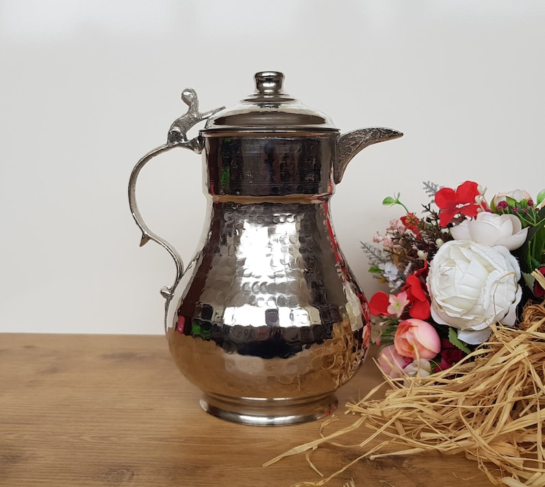 May include: A silver hammered metal pitcher with a decorative handle and lid. The pitcher has a hammered texture and a shiny finish.