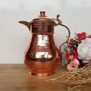 May include: A hammered copper pitcher with a brass handle and spout. The pitcher has a traditional design and is perfect for serving beverages.