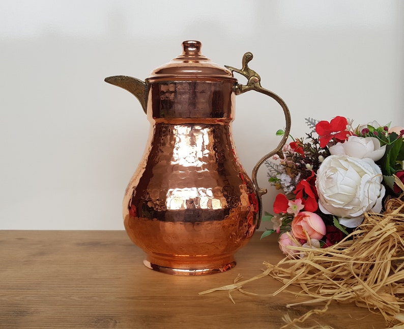 May include: A hammered copper pitcher with a brass handle and spout. The pitcher has a traditional design and is perfect for serving beverages.