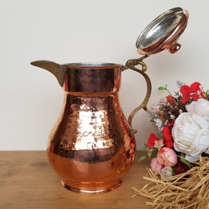 May include: A hammered copper pitcher with a hinged lid and a brass handle. The pitcher is sitting on a wooden surface with a bouquet of flowers in the background.