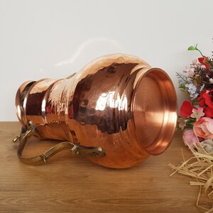 May include: A hammered copper pitcher with a brass handle. The pitcher is on a wooden surface and has a shiny, metallic finish.