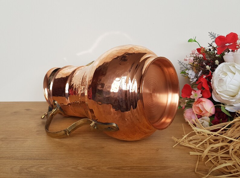 May include: A hammered copper pitcher with a brass handle. The pitcher is on a wooden surface and has a shiny, metallic finish.