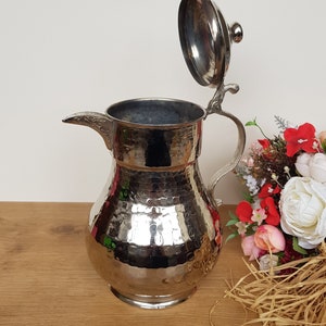 May include: A silver hammered metal pitcher with a hinged lid. The pitcher has a rounded body and a spout. The lid has a small knob on top. The pitcher is sitting on a wooden surface.