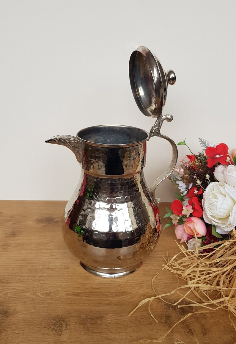 May include: A silver hammered metal pitcher with a hinged lid. The pitcher has a rounded body and a spout. The lid has a small knob on top. The pitcher is sitting on a wooden surface.
