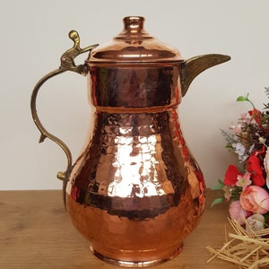 May include: A hammered copper pitcher with a brass handle and spout. The pitcher has a rounded body and a domed lid.