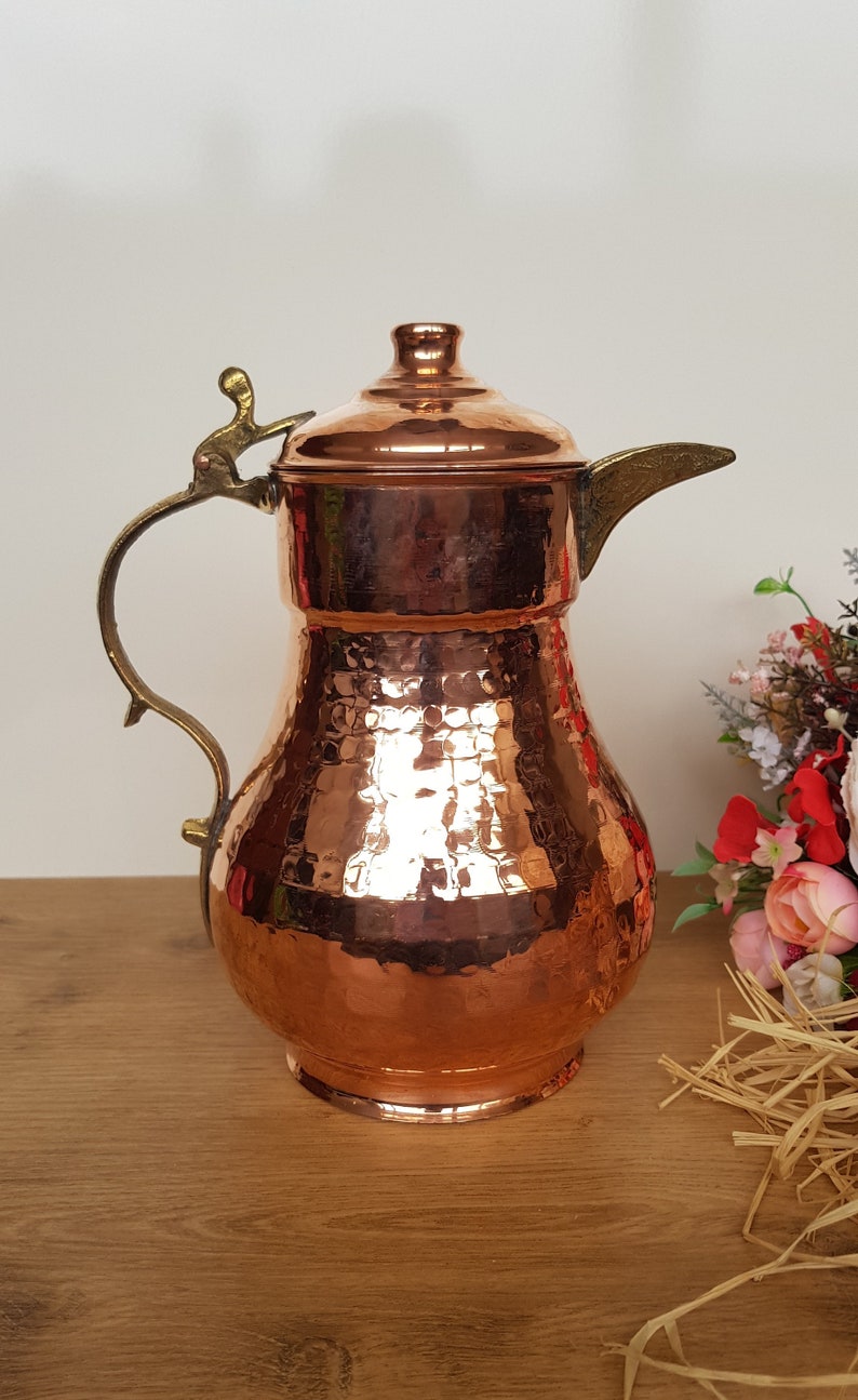 May include: A hammered copper pitcher with a brass handle and spout. The pitcher has a rounded body and a domed lid.