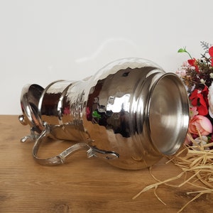 May include: A silver hammered metal pitcher with a lid and a handle. The pitcher is lying on its side on a wooden surface.