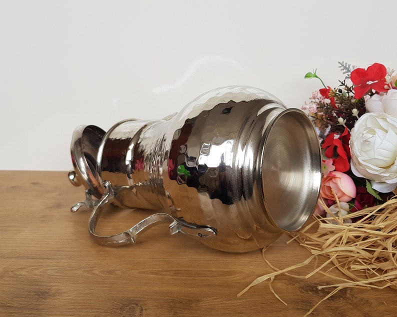 May include: A silver hammered metal pitcher with a lid and a handle. The pitcher is lying on its side on a wooden surface.