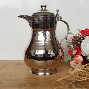 May include: A silver hammered metal pitcher with a lid and a handle. The pitcher is sitting on a wooden surface with a bouquet of flowers in the background.