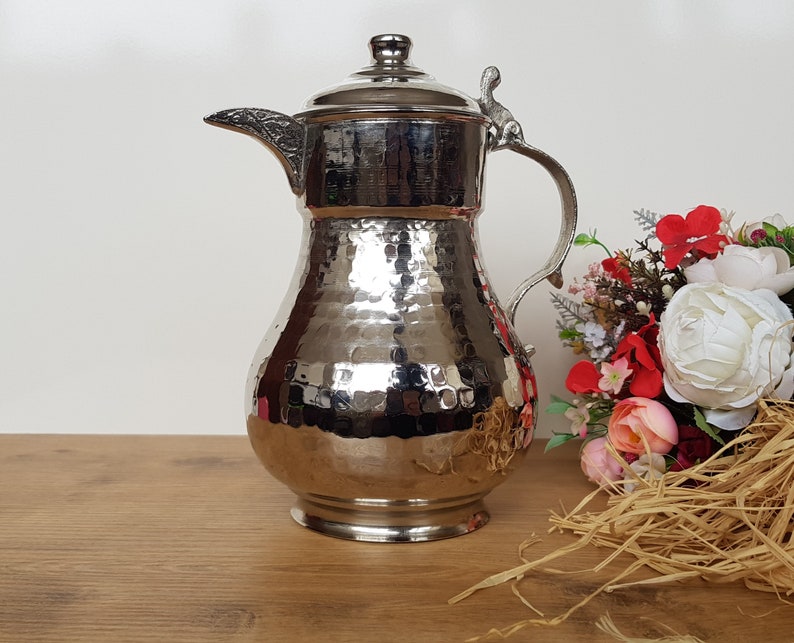 May include: A silver hammered metal pitcher with a lid and a handle. The pitcher is sitting on a wooden surface with a bouquet of flowers in the background.