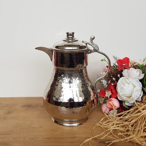 May include: A silver hammered metal pitcher with a spout and a handle. The pitcher has a decorative design and is sitting on a wooden surface.