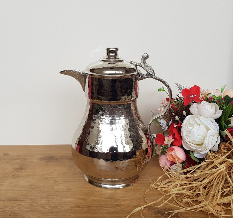May include: A silver hammered metal pitcher with a spout and a handle. The pitcher has a decorative design and is sitting on a wooden surface.