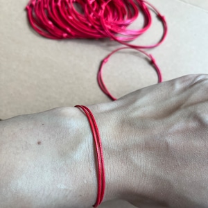May include: A close-up of a red string bracelet on a person's wrist. The bracelet is made of a thin, braided cord and is tied in a simple knot.
