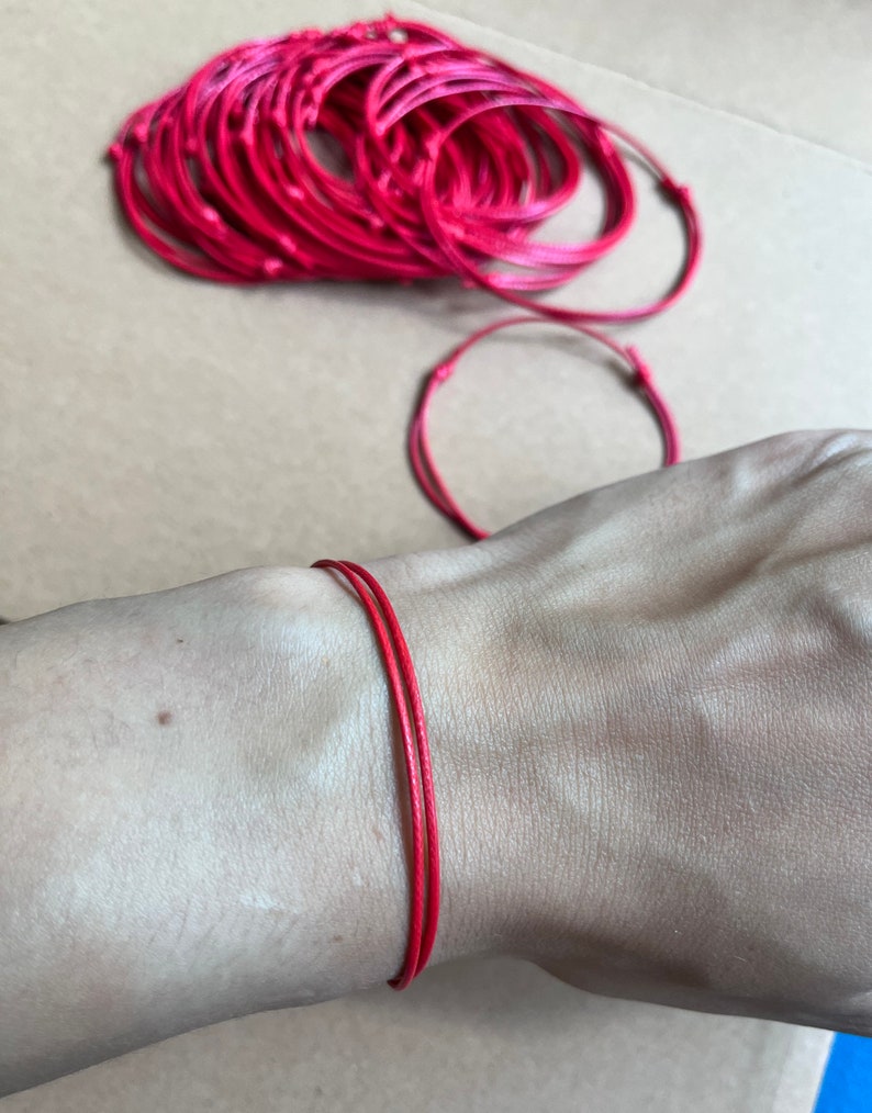 May include: A close-up of a red string bracelet on a person's wrist. The bracelet is made of a thin, braided cord and is tied in a simple knot.