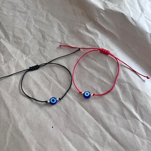 May include: Two adjustable cord bracelets. One is black, the other is red. Each bracelet features a blue evil eye charm with silver accents. The bracelets are displayed on a textured, light brown surface.