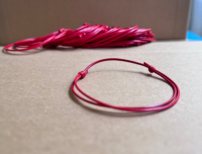 May include: A red cord bracelet with a simple knot closure. The bracelet is made of a thin, flexible cord and is approximately 7 inches in length.