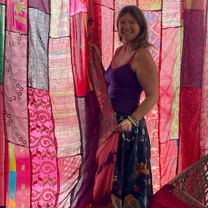 May include: A patchwork curtain made of various fabrics in shades of pink, red, and purple. The curtain is hanging in front of a person wearing a purple top and a patterned skirt. The fabrics have different patterns and textures.