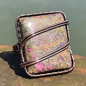 Könnte beinhalten: Handgefertigter Ring mit einem rechteckigen Stein mit einem gesprenkelten Muster in Grün, Braun und Rosa. Der Stein ist in einem kupferfarbenen Drahtrahmen eingefasst, was ein einzigartiges Design ergibt. Der Ring ist ein Schmuckstück.